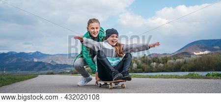 Laughing Kids Have Fun Using A Skateboard. Brother Pushing His Sister Sitting On A Skateboard And Ma