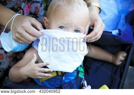 Blond Blue Eyes Baby Looks Straight. Female Hands With Surgery Mask Put White Scarf As A Mask On Chi