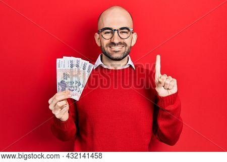 Young bald man holding icelandic krona banknotes smiling with an idea or question pointing finger with happy face, number one 