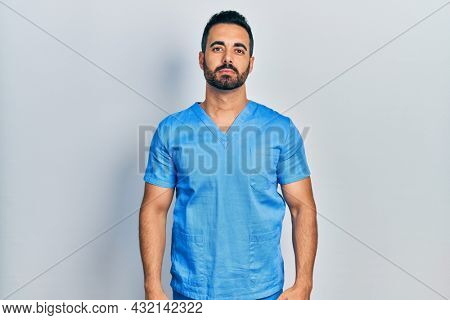 Handsome hispanic man with beard wearing blue male nurse uniform relaxed with serious expression on face. simple and natural looking at the camera. 