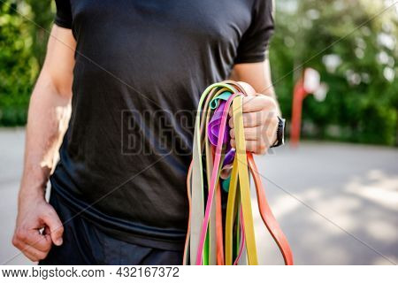 Strong man guy holding in his hand wearing fitness tracker set of colorful elastic rubber bands at the stadium outdoors. Athlete male person during workout with additional sport equipmant