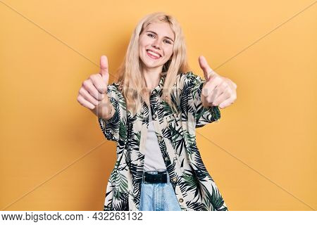 Beautiful caucasian woman with blond hair wearing tropical shirt approving doing positive gesture with hand, thumbs up smiling and happy for success. winner gesture. 