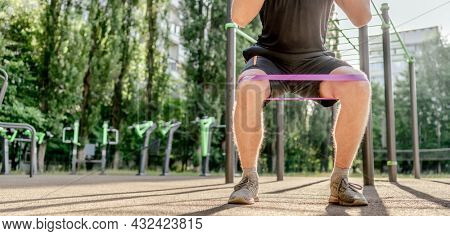 Strong sportive man guy doing squats with elastic rubber band at the stadium outdoors. Adult male person during workout for legs with additional sport equipmant. Sportsman exercising outside