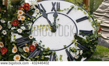 Large Wall Clock On The Field In Blooming Flowers, Showing The Time 11:55, 12:00. Time Management Co