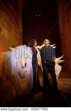Couple In Old Dark Clothes In Abandoned Castle With Old Brick Walls. Man In Black Dress And Woman Wi