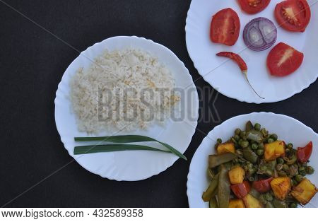 Closeup High Angle View Of Rice, Matar Paneer Mix Veg And Salad On White Plate Over Black Background
