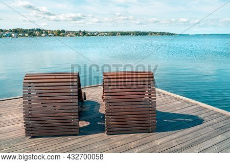 Two Wooden Beach Beds Or Chairs On A Wooden Pier Next To The Sea With Cityscape In The Background. S