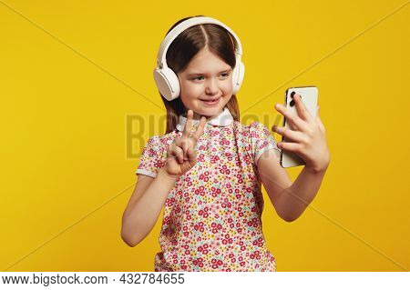 Caucasian Girl Shows Peace Gesture, Uses Smartphone, Listens Favorite Music Via White Headphones Wea