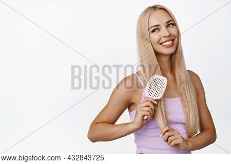 Good-looking Young Blond Woman Combing Hair, Holding Comb Brush And Smiling, Looking Aside, Standing