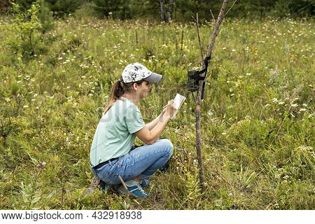 Young Woman Scientist Zoologist Writing Down Data From Trap Camera To Notepad, Observing Wild Animal