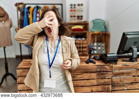 Young down syndrome woman working as manager at retail boutique peeking in shock covering face and eyes with hand, looking through fingers with embarrassed expression. 