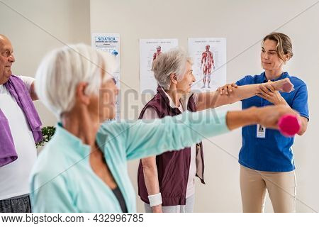 Group of old people using dumbbells for workout session with physiotherapist. Group of active seniors exercising in clinic with trainer. Elderly woman lifting weight for wellness with trainer.