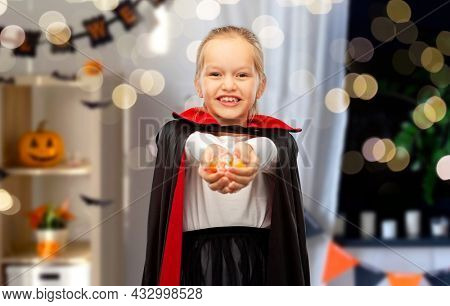 halloween, holiday and childhood concept - happy smiling girl in black dracula cape or costume with candies over decorated home room and lights background