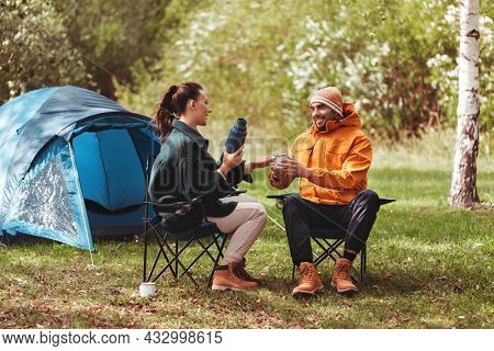 camping, tourism and travel concept - happy couple with thermos drinking tea at tent camp