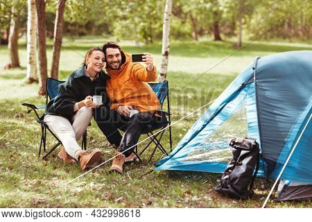 camping, tourism and travel concept - happy couple with smartphone drinking tea and taking selfie at tent camp