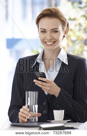 Portrait of happy young businesswoman using mobilephone, drinking water and coffee.