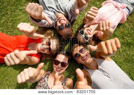 friendship, leisure, summer, gesture and people concept - group of smiling friends lying on grass in circle and showing thumbs up outdoors