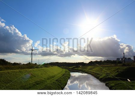 A wind turbine in a peaceful landscape with green grass blue skye and a river