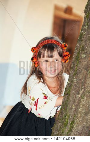 Smiling little girl in the national Ukrainian Hutsul costume and in a wreath looks out from behind a tree