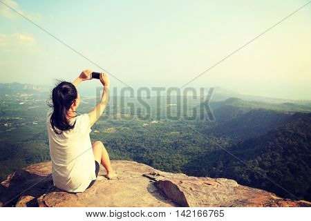 young asian woman taking photo with smart phone at mountain peak cliff