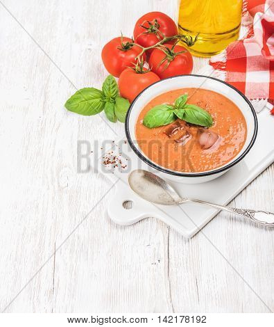 Cold gazpacho soup in bowl with ice, hot pepper and basil served with fresh tomotoes on ceramic board over white wooden background, top view, selective focus, copy space, vertical composition