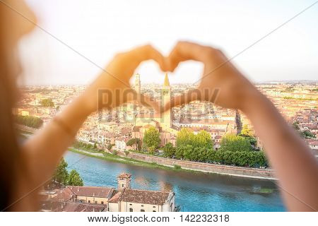Female hands making heart shape on Verona cityscape background. Verona is famous city of love in the north of Italy.