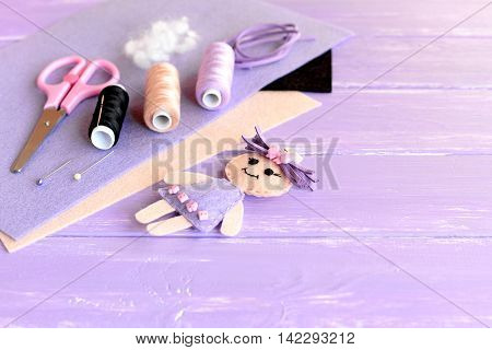 Funny doll decorated with buttons, thread set, needle, pins, scissors, flat pieces of felt on wooden background with blank place for text. Supplies for sewing plush mini doll. Easy hand sewing for kid