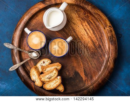 Coffee espresso in cups with cookies and milk in jug on wooden serving round board over dark blue painted plywood background, top view, copy space