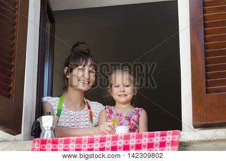 Two years old cheerful daughter with her drinking coffee mother are sitting near open window with traditional European wood brown shutters