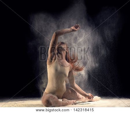 Woman dancer lying on the floor in a ballet pose with flour. Redheaded woman ballerina posing in beige bathing suit on a black background.