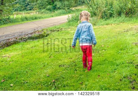 Little toddler girl with blonde pigtails en red boots walking through the wet grass. Her pants are dirt from splashing in the puddles.