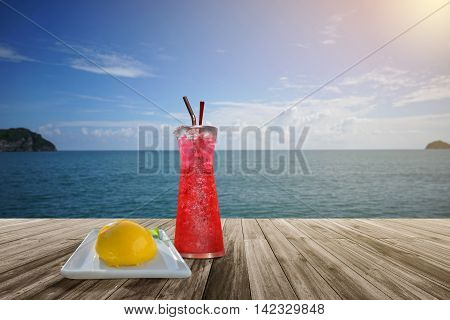 red soda and orange sponge cake on Wooden table front of blurred background. Perspective brown wood over blur sea and blue sky with cloud summer season. sunlight effect on right side of picture