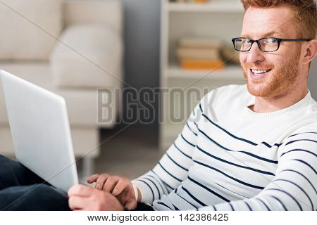 Modern generation. Cheerful handsome smiling man using laptop and looking at you while sitting at the table