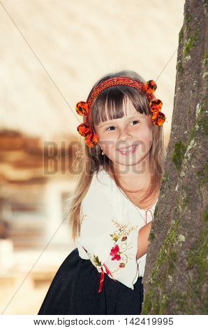 Smiling little girl in the national Ukrainian Hutsul costume and in a wreath looks out from behind a tree