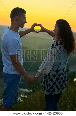 romantic couple at sunset show a heart shape from hands, beautiful landscape and bright yellow sky, love tenderness concept, young adult people