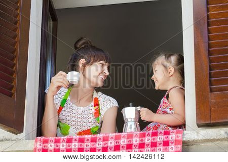 Laughing and looking at each other mother with two years old daughter are holding traditional Italian coffee set of white cup and metal moka pot