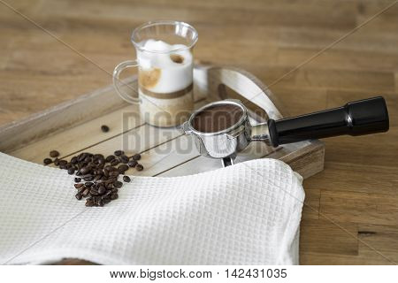 Glass of latte macchiato, coffee beans and a grouphead on a wooden tray