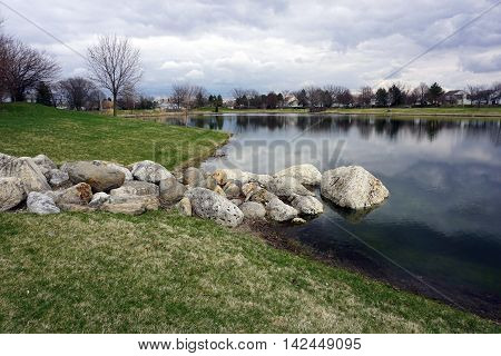 A pile of large rocks on the shore of a small, man-made lake in the Wesmere Country Club subdivision of Joliet, Illinois during the Spring.