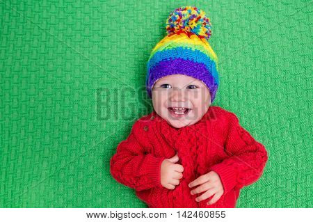 Cute baby in warm wool knitted hat on a red blanket. Autumn and winter clothing for young kids. Colorful knitwear for children. Adorable little boy ready for a walk on a cold fall day.