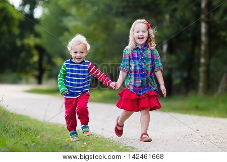 Kids playing in autumn park. Children play outdoors on a sunny fall day. Boy and girl running together hand in hand in a forest. Toddler and preschooler pick colorful oak leaf. Family fun outdoor.