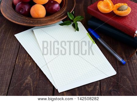 White sheets of paper for your text. Books for records and a pen in the foregroud. Ripe plums and apricots in a clay bowl in the background.
