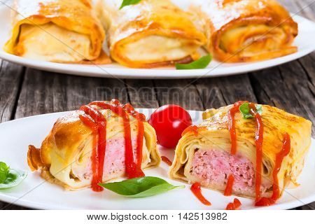 rolled pancakes or crepes stuffed with minced meat poured with tomato sauce on oval dishes served with cherry tomatoes and basil view from above close-up