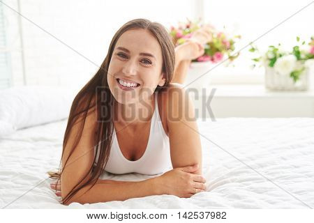 An attractive young Caucasian woman on the bed is radiant with the joyful emotions while lying on the bed and crossing her legs
