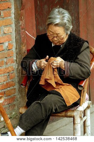 Pengzhou China - April 13 2006: Elderly Chinese woman cutting a piece of fabric sitting on a bamboo chair in front of her home on Hua Lu (Old Street)