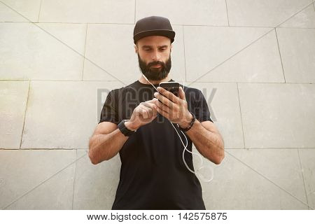 Bearded Muscular Man Wearing Black Tshirt Blank Snapback Cap Summer Time.Young Men Standing Opposite Empty Gray Concrete Wall Background Using Smartphone Headphones.Horizontal Mockup
