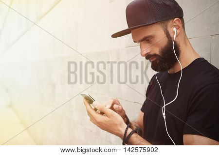 Bearded Muscular Man Wearing Black Tshirt Blank Snapback Cap Summer Time.Young Men Standing Opposite Empty Gray Concrete Wall Background Using Smartphone Headphones.Horizontal Mockup.Side View