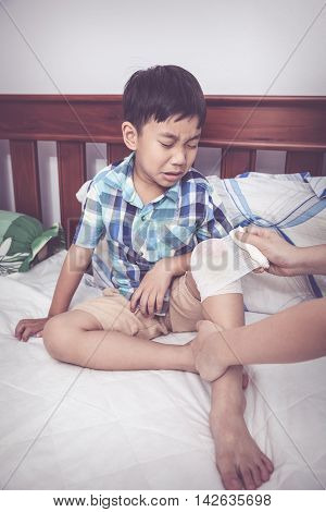 Crying child injured. Mother bandaging son's knee on bed inside bedroom bandage in focus asian boy sadden and crying. Human health care and medicine concept. Vintage tone effect.