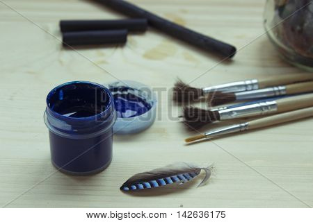 Art tools still life with brushes blue gouache paint charcoal and a feather on a wooden table. Selective focus.