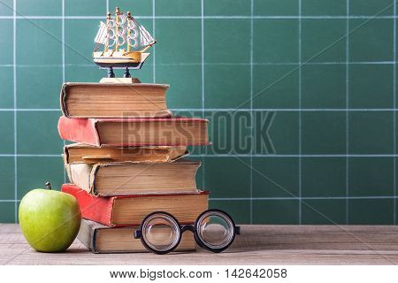 The old school books textbooks and supplies lie on a wooden table. Tutorials on a green blackboard in cage background.