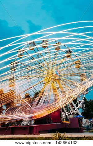 Motion Blurred Effect Around Of High Speed Rotating Illuminated Attraction Feature In City Amusement Park. Ferris Wheel On Summer Evening Blue Sky Background.